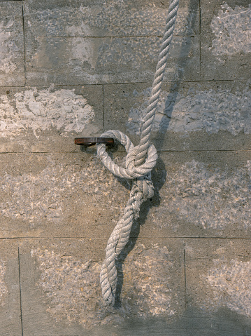 White thick rope attached to a wall rusty metal ring by a knot against a stone brick wall direct view