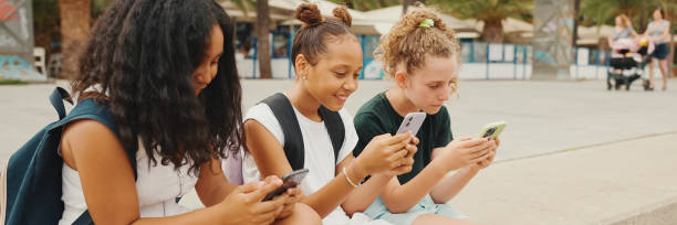 três amigas pré-adolescentes estão sentadas na orla usando celular. três adolescentes jogando em um smartphone ao ar livre em fundo urbano - mobile phone group of people human face latin american and hispanic ethnicity - fotografias e filmes do acervo