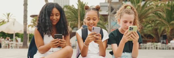 três amigas pré-adolescentes estão sentadas na orla usando celular. três adolescentes jogando em um smartphone ao ar livre em fundo urbano - mobile phone group of people human face latin american and hispanic ethnicity - fotografias e filmes do acervo