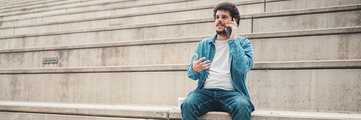 young bearded man in denim shirt sitting on high steps and using mobile phone. Man talking on smartphone