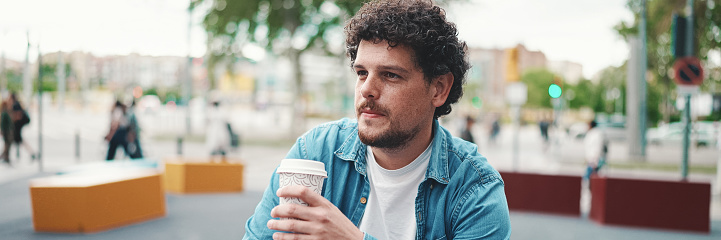 Close-up portrait of young bearded man in denim shirt sitting in wireless headphones and drinking coffee on busy street modern city background. Profile of man drinking coffee