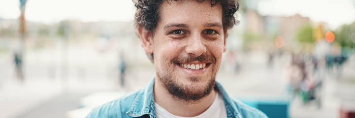 closeup portrait of young bearded man in denim shirt posing and smiling at camera on modern cityscape background.