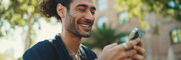 istock Attractive italian guy with ponytail and stubble is using mobile phone sitting on bench at old buildings background. Stylish man writes message, views photo on smartphone 1733716977
