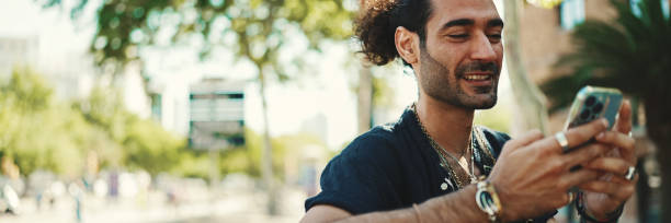 istock Attractive italian guy with ponytail and stubble is using mobile phone sitting on bench at old buildings background. Stylish man writes message, views photo on smartphone 1733723125