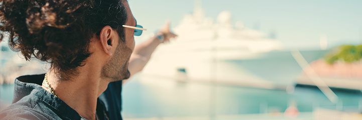 Clouse-up,young italian guy with long curly hair and stubble takes selfie on mobile phone. Stylish man in sunglasses posing smiling at camera of his smartphone and pointing at yacht moored in port