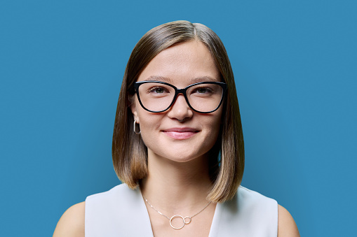Headshot portrait of young beautiful woman in glasses, female face with natural trendy makeup hairstyle on blue studio background. Beauty care skin hair, business education fashion, health vision eyes