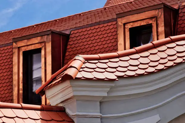 Sloped red brown tile Mansard roof closeup. shiny red copper plated dormers. beavertail tiles. entrance gate detail. white stucco elevation. modern materials. residential building.