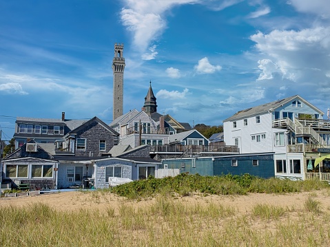 Old town Provincetown from the beach looking back at cottages and Pilgrim monument. Sand dunes, to commercial district to the highest points with church steeple and Pilgrim monument in distance.