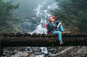 Young woman with backpack sitting on a wooden bridge and enjoying of power mountain river waterfall during Makalu Barun National Park trek in Nepal. Mountain hiking and active people concept image.