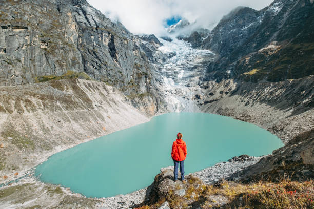 Female trekker dressed bright red jacket on the rock enjoying a glacier falling in high altitude Sabai Tso glacial lake cca 4350m. Makalu Barun National Park, Mera peak climbing route, Nepal.