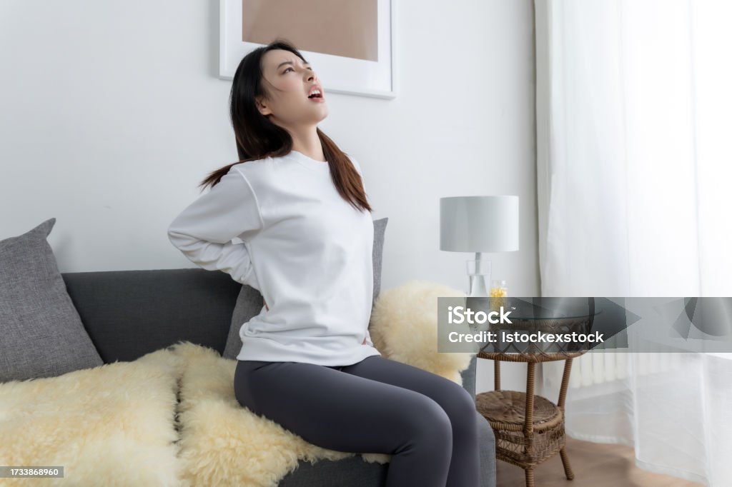 Young pretty Asian girl sitting on a cozy sofa in the living room and with back pain. Backache Stock Photo