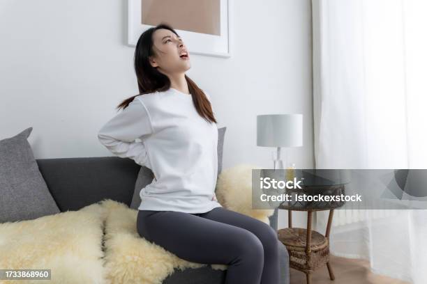 Young Pretty Asian Girl Sitting On A Cozy Sofa In The Living Room And With Back Pain Stock Photo - Download Image Now