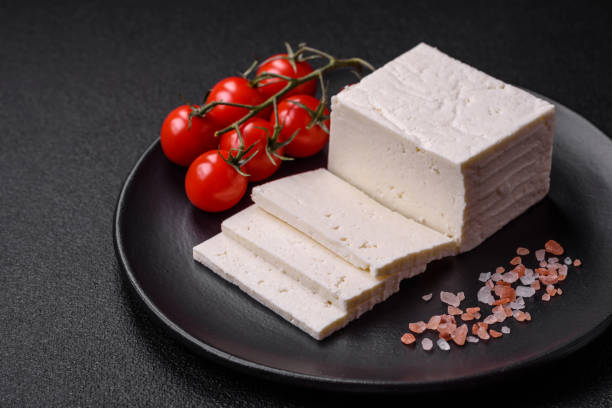 Delicious fresh ricotta cheese cut into slices with vegetables, salt and spices Delicious fresh ricotta cheese cut into slices with vegetables, salt and spices on a dark concrete background feta cheese block stock pictures, royalty-free photos & images