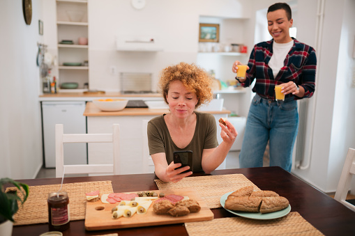 Beautiful woman using a smart phone at breakfast table with her girlfriend bringing her a glass of orange juice in kitchen. Lesbian couple having breakfast in kitchen at home.