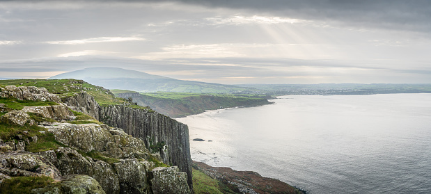 Wide panoramic view of rocky cliffs and shoreline at Fairhead, County Antrim, Northern Ireland. Ballycastle town is in the distance and the sky is cloudy with sun beams shining through. The sea is calm and season is autumn. The landscape was used in TV series Game of Thrones and is popular area for tourism.