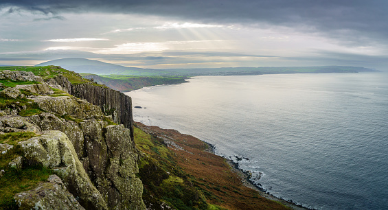 Wide panoramic view of tall cliffs and the shore at Fairhead, County Antrim, Northern Ireland. Ballycastle town is in the distance and the sky is dark and moody, cloudy with sun beams shining through. The sea is calm and season is autumn. The landscape was used in TV series Game of Thrones and is popular area for tourism.