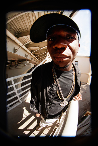 Fish eye shot of African American guy in stylish casualwear looking at camera while standing by railings in urban environment on sunny day