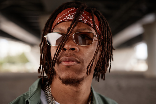 Face of young stylish African American male rapper or hip hop dancer in headband and sunglasses standing in front of camera outdoors
