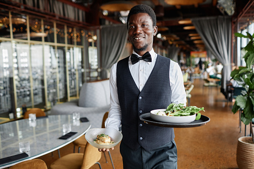Portrait of young Black man as server bringing food on tray to guests in restaurant and smiling happily