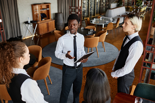 High angle portrait of restaurant manager talking to servers wearing classic uniforms during staff meeting in modern dining room