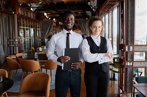 Waist up portrait of two young people as luxury restaurant staff wearing classic black and white uniforms smiling at camera, copy space
