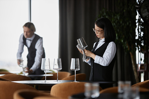 Side view portrait of young Asian woman polishing wine glass in luxury restaurant setting, copy space
