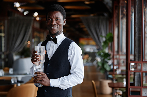 Waist up portrait of young Black man as elegant server holding glasses and smiling at camera in luxury restaurant, copy space