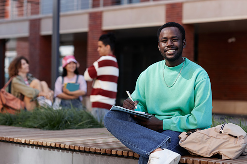Full length portrait of young Black man as student doing homework outdoors on college campus and smiling at camera, copy space
