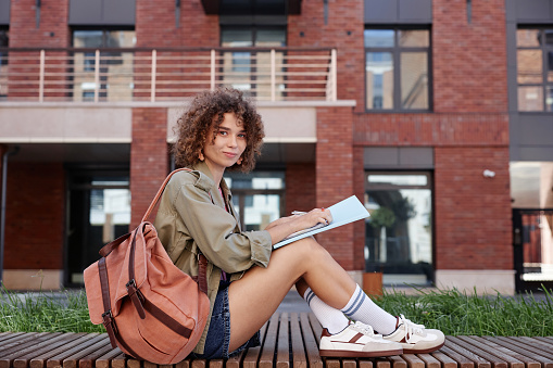 Side view portrait of cute young woman as female student sitting outdoors on college campus and smiling at camera while doing homework, copy space