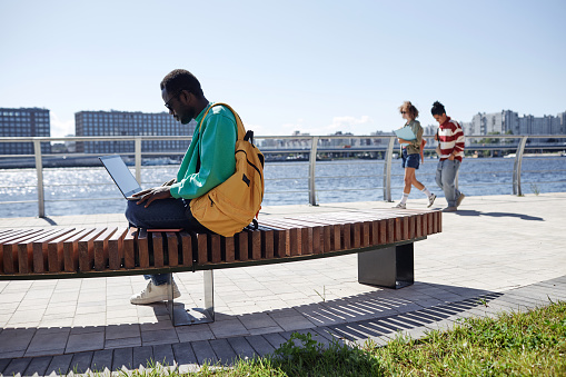 Side view portrait of Black young man as student using laptop outdoors relaxing on bench in riverside park at college campus, copy space