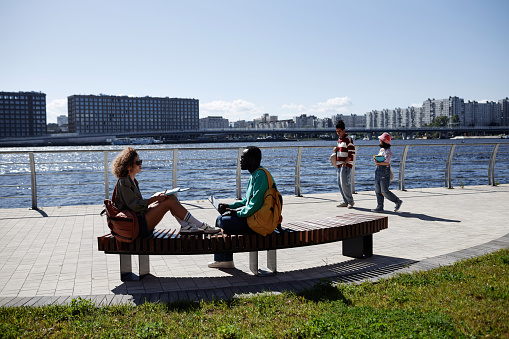 Wide angle view at group of young people relaxing in riverside park outdoors with blue sky and skyline, copy space