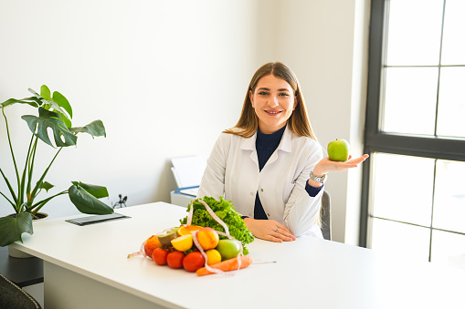 Blonde female healthy nutritionist with fruits and vegetables on her desk in the office shows an apple in her hand