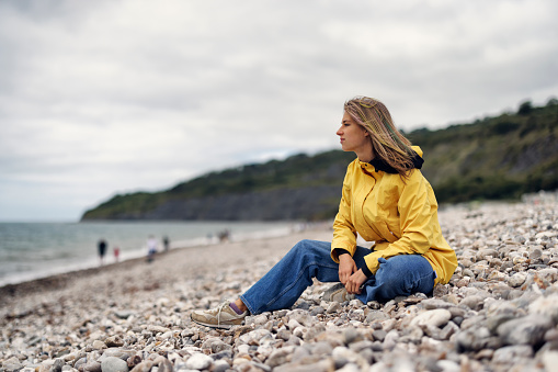 Teenage girl enjoying vacations in Dorset, United Kingdom. \nShe is sitting on the Lyme Regis Fossil Beach.\nCold and windy overcast day.\nShot with Canon R5