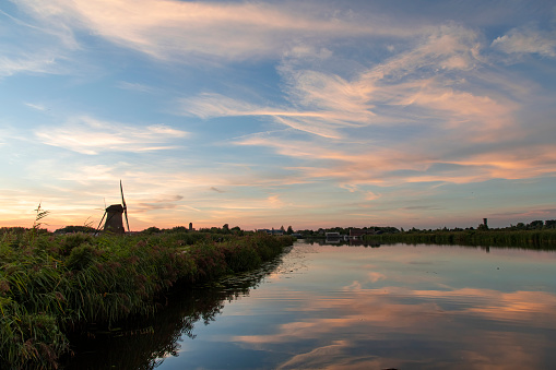 Sunset view over tranquil water with reed beds of a canal with perfect reflection of sunset clouds in water next to silhouette of iconic 18th-century windmill in Kinderdijk, the Netherlands