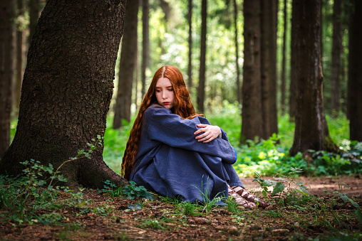 Red haired young woman in a linen blue dress is sitting under a tree of the forest in the rays of the sun. The girl experiences emotions of despair left alone in the forest.