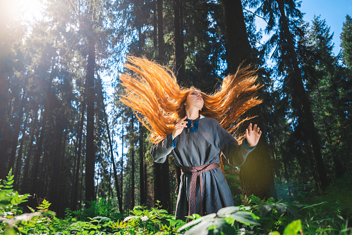 Red haired young woman in a linen blue dress is sitting in the middle of the forest in the rays of the sun. The girl experiences emotions of despair left alone in the forest.