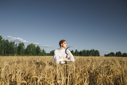 Slavic girl in a white dress and a black skirt with witch makeup stands in a field of rye and holds a terrible sickle in her hands. The concept of a mystical harvest.