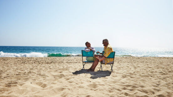 A laughing senior couple sitting in beach chairs with her legs stretched across his on the beach in Santa Monica, California...