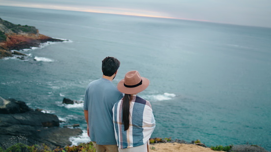 Romantic young couple walking to beautiful sea shore back view. Relaxed tourists going down hill to amazing cloudy seascape. Calm family pair looking on picturesque ocean. Vacation honeymoon concept.