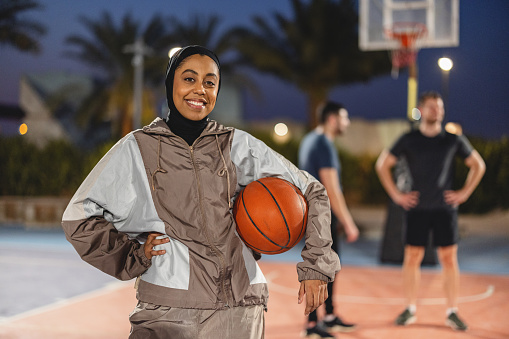Portrait Young Adult Female Athlete  With A Black Hijab Holding A Basket Ball With One Hand And The Other Hand Is On Her Hip. She Is Smiling Showing Her Teeth. She Is Looking At The Camera Outdoors At Night In Dubai.