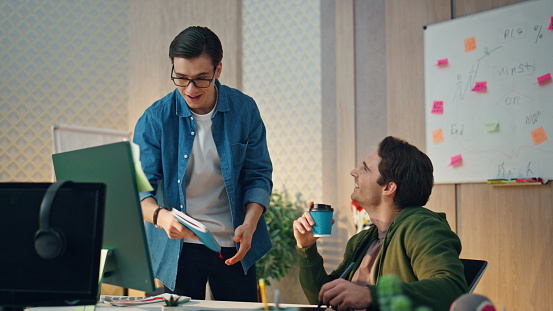 Smiling colleagues working computer in corporate office. Two creators looking pc enjoying friendly conversation. Eyeglasses man saying joke going to break. Laughing guy drinking coffee feeling relaxed