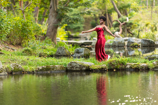 A Black woman in red dress joyfully walking through a Japanese public park by a lake.