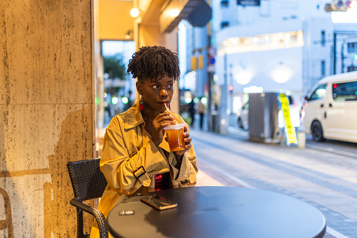 A dark skin woman in formal wear sits at an outdoor cafe seat and table, watching the busy city life go by while she sips on her ice tea.