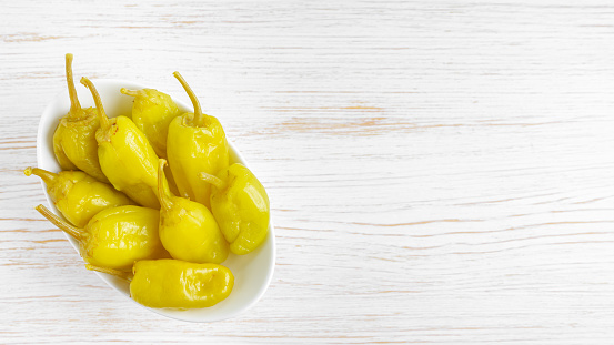 Pickled yellow pepper, pepperoncini or friggitelli in bowl on white wooden background. Hot pepper marinated, brined. Traditional Italian and greek cuisine, ingredient for salad, pasta, sauce.
