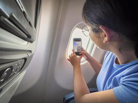 A Eurasian young woman takes a smartphone video through the window of the airplane wing and aerial view of the landscape below.  Denpasar, Bali, Indonesia.