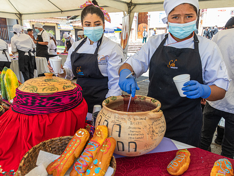 Cuenca, Ecuador-October 30,2022: Young women serve Guagua bun and drink Colada Morada. The bread is molded in a shape of baby. This is the traditional bun and drink used at the Day of the Dead event.