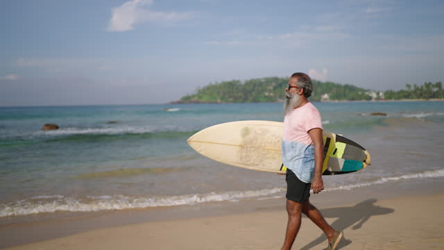 Senior traveler surfer holds surf board and walking at exotic beach on tropical island. Black old bearded man in sunglasses on retirement enjoy tourism, view of ocean, sky, sunlight and rocky hills.