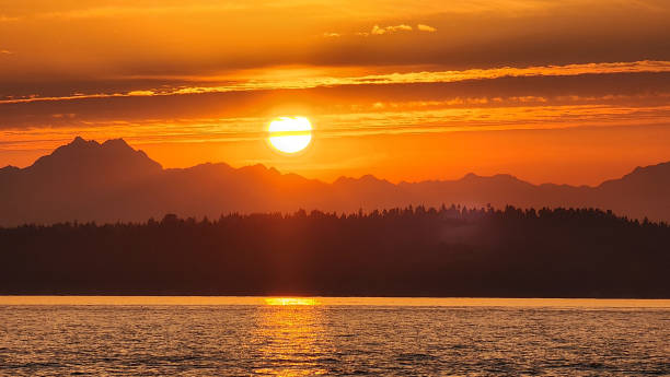A vibrant orange sunset taken from the Puget Sound near Seattle Washington. The bright Golden Sun sets behind the majestic Cadscade Mountain Range and the light reflects off the water of the sea.