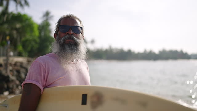 Senior male traveler walking on beach with surfboard, has active and healthy lifestyle in golden years, looking around, enjoying views of bay with boats, exotic nature and plants, ocean with waves.