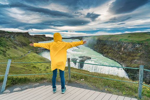 Female tourist in yellow jacket enjoying on viewpoint of Gullfoss waterfall or Golden Falls with extreme hvita river flowing in canyon on summer at Iceland